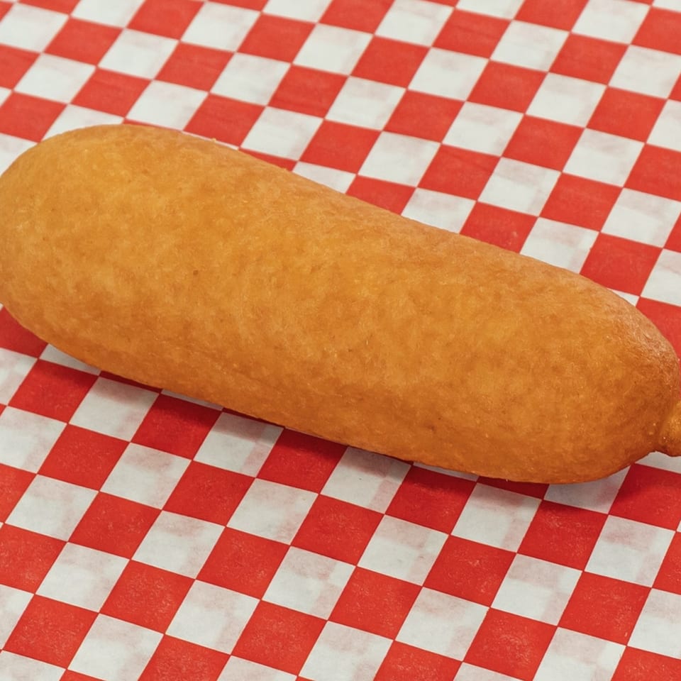 A golden-brown corn dog rests diagonally on a red and white checkered paper liner.