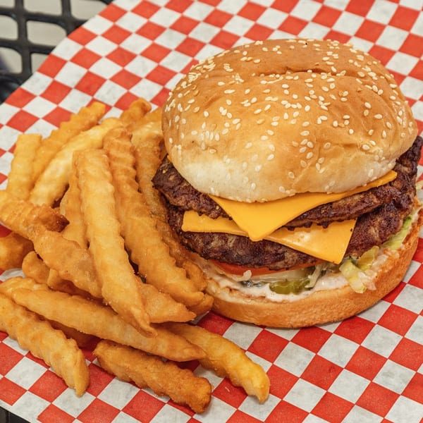 A double cheeseburger with lettuce, tomato, and pickles, served with crinkle-cut fries on checkered paper.