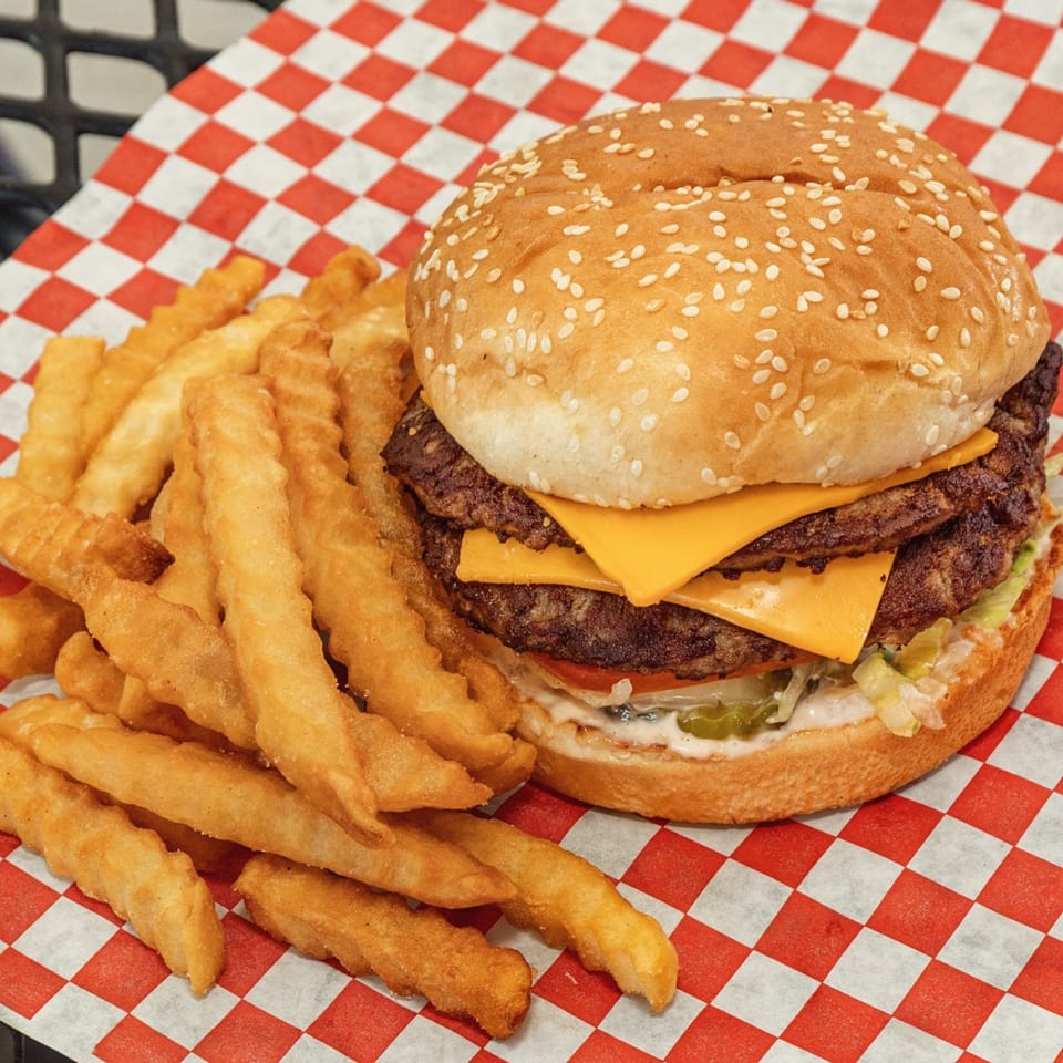 A double cheeseburger with lettuce, tomato, and pickles, served with crinkle-cut fries on checkered paper.