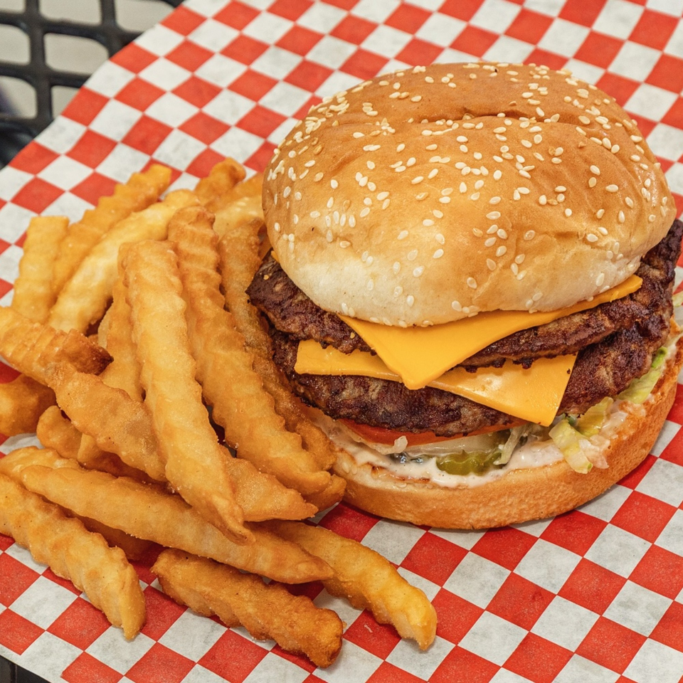 A double cheeseburger with lettuce, tomato, and sauce, served with crinkle-cut fries on red and white checkered paper.